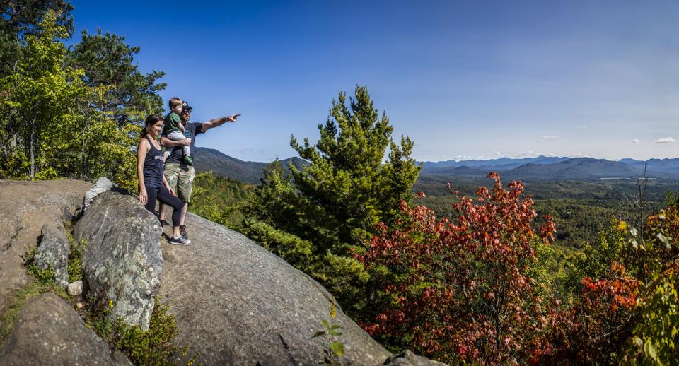 A couple holding a toddler points off into the distance from the top of Baker Mountain with some of the trees closest to them starting to turn red