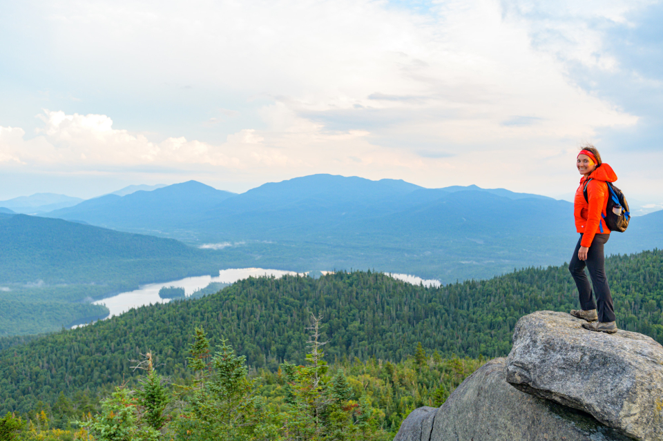 A female hiker smiles from a rocky pinnacle with mountain views beyond.