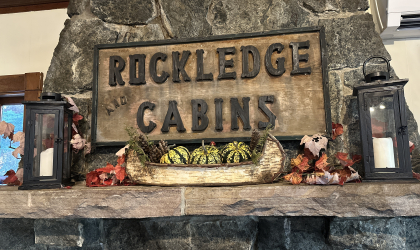 A carved sign reading 'Rockledge and Cabins' over a stone mantle with birch bark boat filled with festive squash and candles.