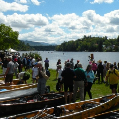 Large group of people wait on the green by the water with their canoes