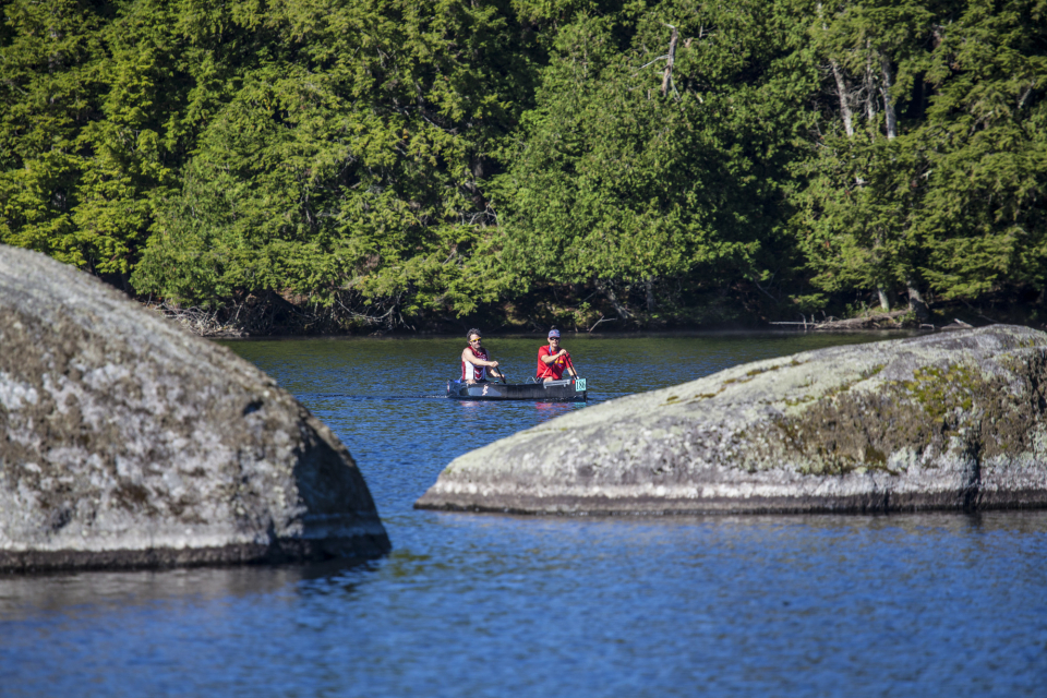 Two people paddling a blue canoe on a calm lake with trees in the background. Two large rocks poke through the water in the foreground.