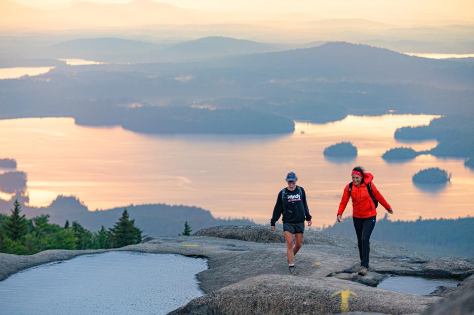 Two hikers atop a rocky summit with an expansive lake and forested islands in the background.