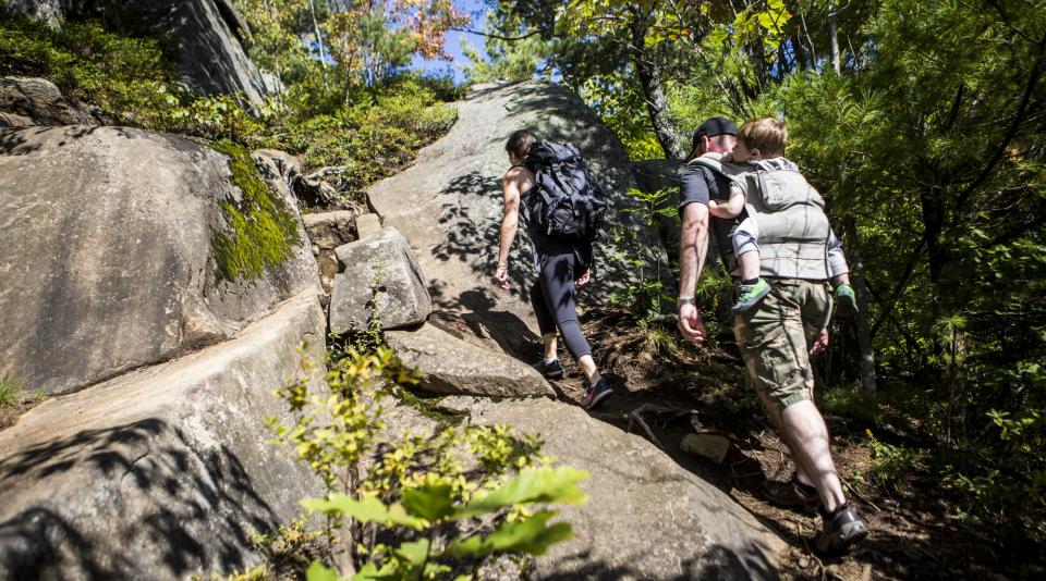 Two hikers, one carrying a child in a backpack, hike up a rocky, tree-lined trail.