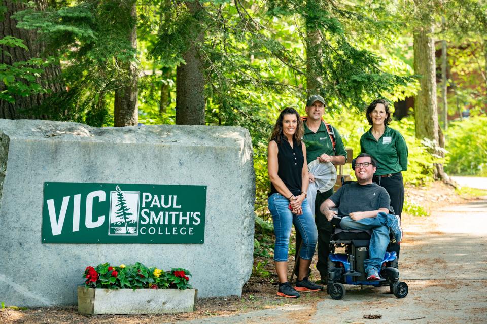 A man in a mobility chair and three attendants from Paul Smith's College pose next to the sign.