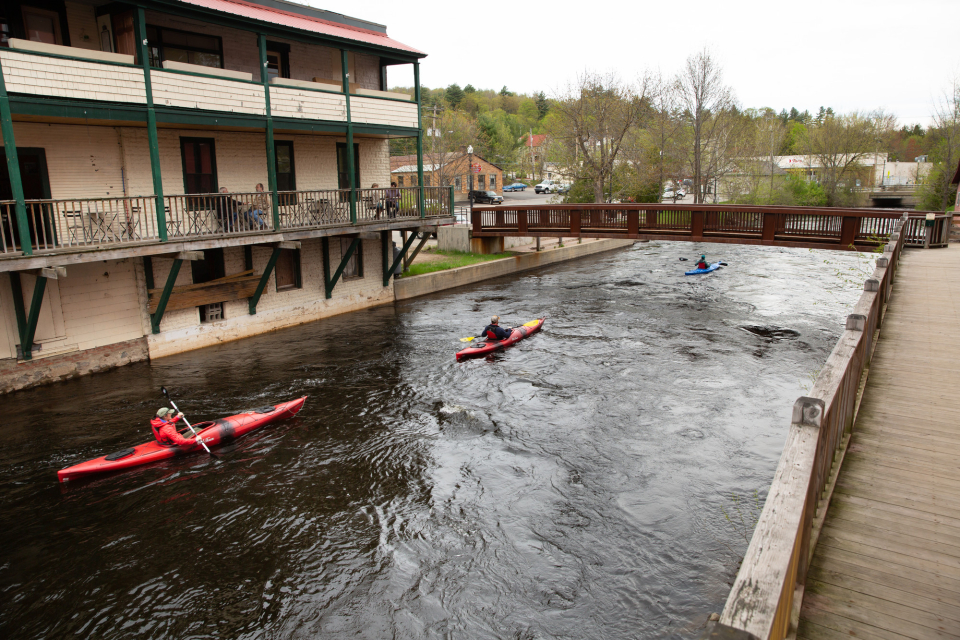 Three kayakers paddling through downtown Saranac Lake on the river as people dine at a cafe nearby.