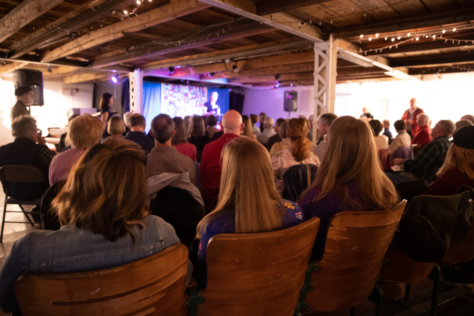 A group of people sit and watch a performance at a rustic art studio.