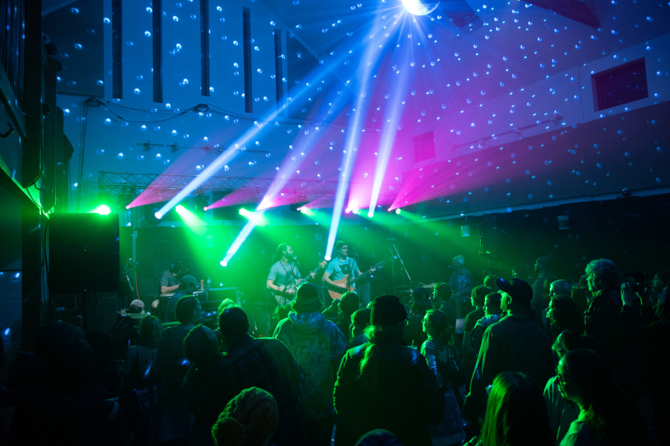 A crowd of people watch a band perform under purple and green lights.