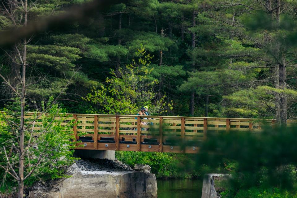 Two people ride bikes over a bridge tucked in the woods.