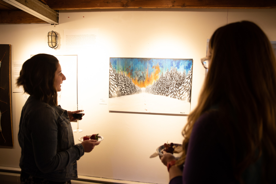 Two individuals admire a vibrant winter landscape painting in a warmly lit art gallery, holding plates of snacks and glasses of wine.