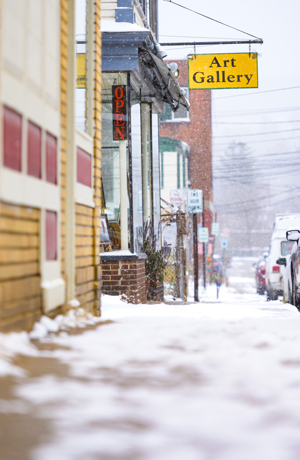 Snowy sidewalk leading to a cozy art gallery in Saranac Lake with a bright yellow sign.