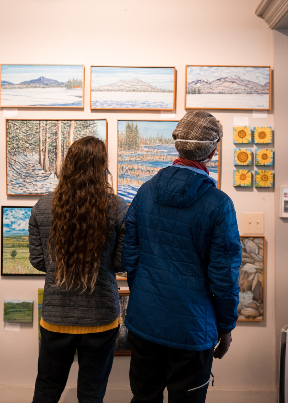 Two people admiring winter landscape paintings in a cozy art gallery.