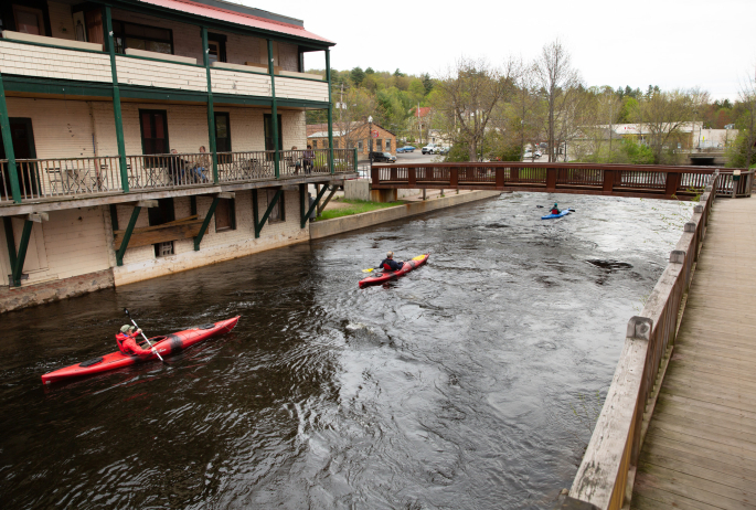 Three kayakers paddling through downtown Saranac Lake on the river as people dine at a cafe nearby.