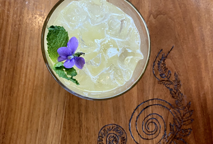 lemonade with flower garnish on table with wood carving of a fiddlehead fern