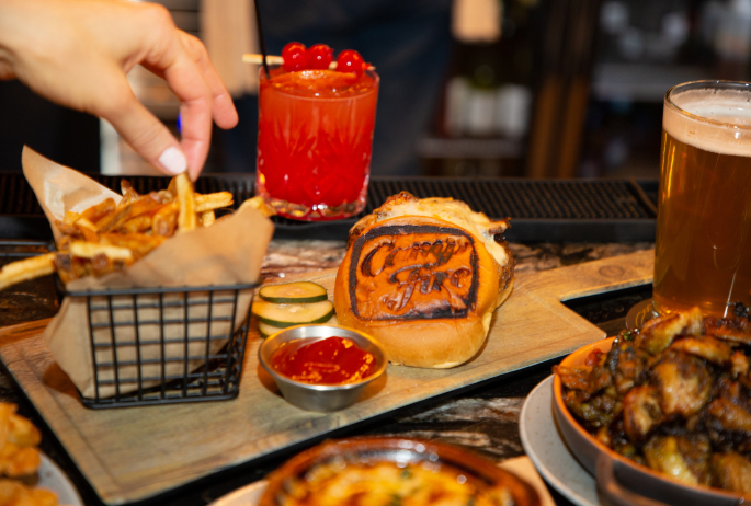 Fries, a cocktail, a beer, and burger with a campfire branding on the bun.