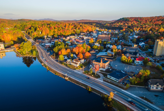 An aerial shot of downtown Saranac Lake. Fall foliage, lakes, buildings, and the highway are featured.