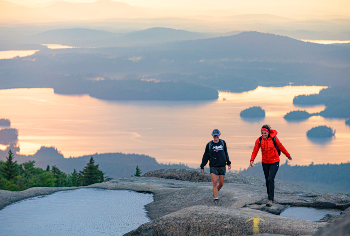 Two people walk along an open rocky summit with a large lake behind them