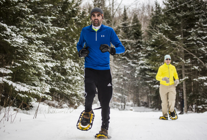 A man and woman run in snowshoes on a snowy trail.