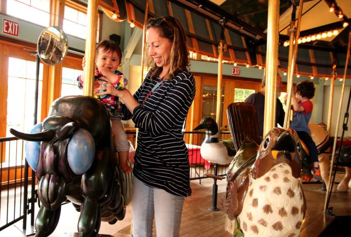 A woman holds her child up on an bug ride on a carousel.