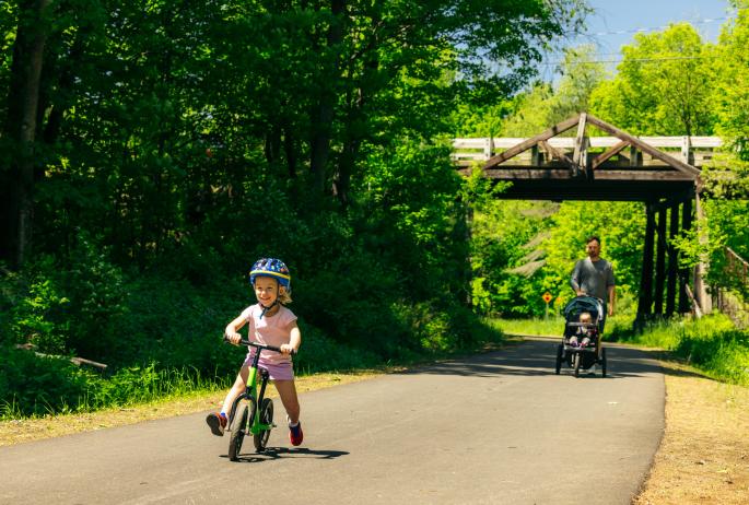 A child rides their tricycle in front of a man pushing a stroller.