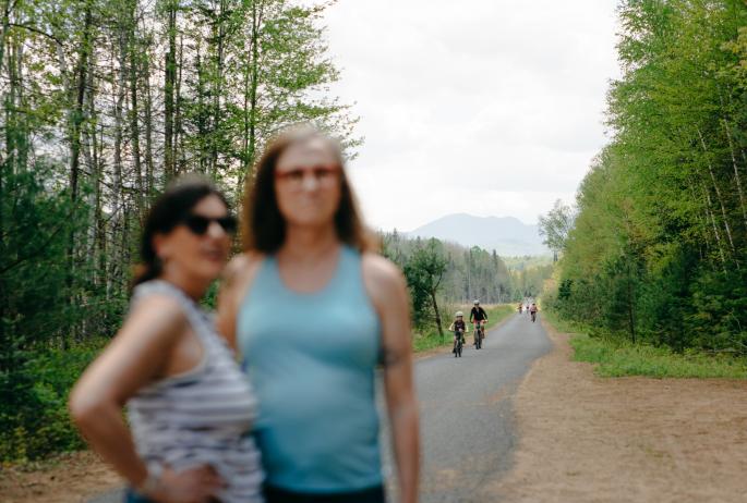 Two women pose for a photo with bicyclists in focus in the background on a trail.