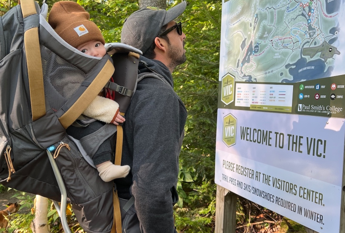 a man reads a sign with a baby strapped to his back.