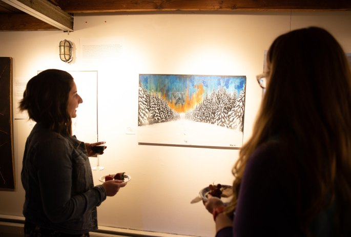 Two individuals admire a vibrant winter landscape painting in a warmly lit art gallery, holding plates of snacks and glasses of wine.