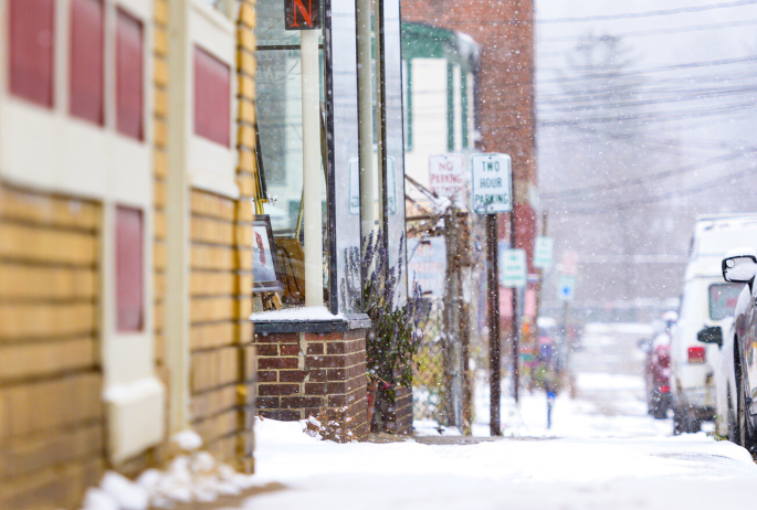 Snowy sidewalk leading to a cozy art gallery in Saranac Lake with a bright yellow sign.