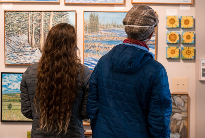 Two people admiring winter landscape paintings in a cozy art gallery.