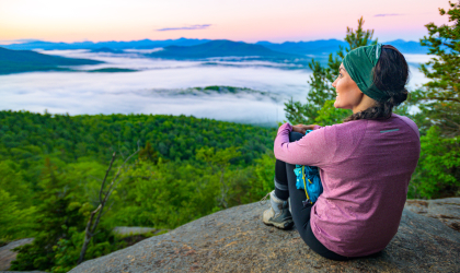 Woman in a purple shirt sits on the edge of Baker Mountain and stares out overtop of the green trees below with light cloud cover and other Adirondack mountains in the distance