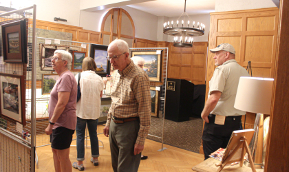 People observe the art inside a large room with wood paneled walls at the guide boat art show