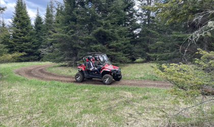 A red UTV sits on a well ridden trail in the grass in front of a large pine tree