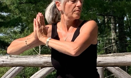 A woman holds a yoga pose with her eyes closed outside surrounded by pine trees.